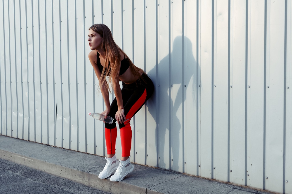 Female athlete drinking water while standing by a grey wall. Slim woman in sportswear taking a break after workout.