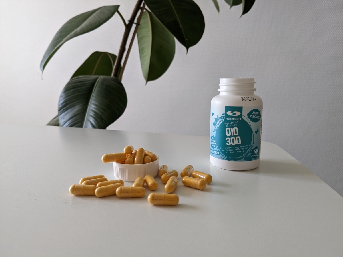 Healthwell Q10 300 fotograferad av oss under testet, burk och kapslar på bord med grön växt i bakgrunden