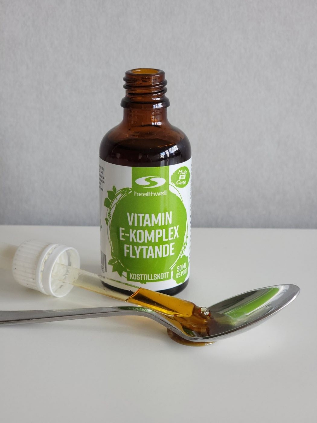 Vår testbild av Healthwell Vitamin E Komplex Flytande med pipett och en gyllene, trögflytande droppe i sked