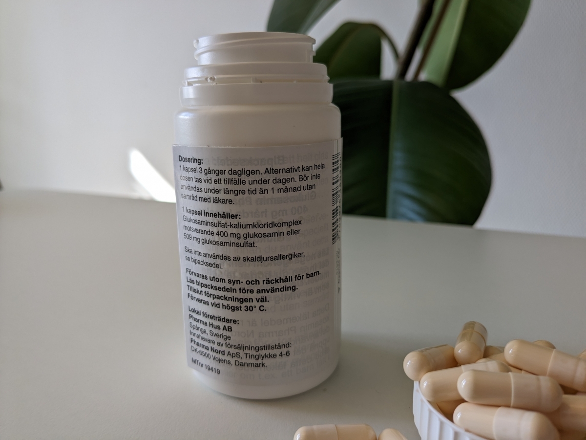 Baksida på Pharma Nord Glukosamin med tydlig doseringstext och kapslar i förgrund, foto av oss
